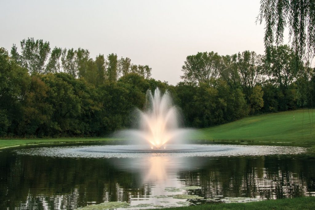 Kasco 8400JF fountain producing tall illuminated 3 tier spray display in large landscaped pond at dusk.
